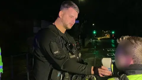 A traffic officer dressed in black uniform holds out a breathalyser device while a young man, whose face is blurred, blows into it on a dark street.