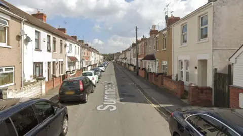 A Google Maps street view of a residential street, with cars parked on either side of the road and a row of terraced houses.