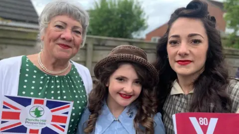 An older woman, a girl and a younger woman stand together smiling. They all have their hair and make-up done in a 1940s style and are also wearing retro dresses. They are holding VE Day and Union Jack flags. 