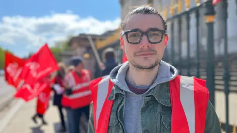 Tom Jackson/BBC David Jackson is looking at the camera and standing in front of railings. He is wearing a hi-vis orange jacket over a pullover. His hair is combine back and he has thick-rimmed spectacles on. In the background are blurred images of people waving red banners on the pavement.