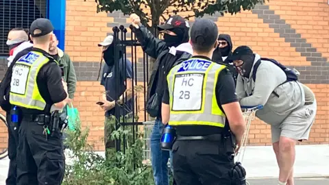 Four men wearing masks, one of whom appears to be shaking his fist in the air, standing behind a line of police