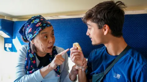 BBC/Studio Lambert An elderly woman holds Kush's hand as she tells him to eat a food item while they on a train in Kazakhstan.