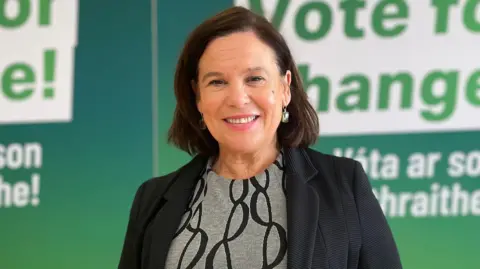 PA Media Mary Lou McDonald, a woman with shoulder-length brown hair, is smiling at the camera as she stands in front of green and white party posters. She is wearing a grey jumper with black circle designs and a black blazer jacket.