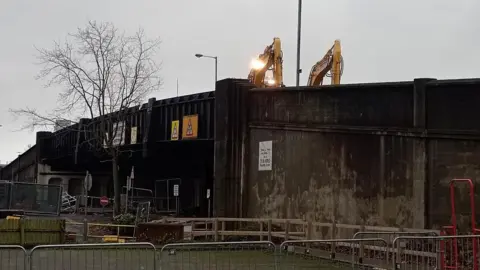 A partial view of the top of two yellow diggers working on the Boyne Bridge in Belfast.   The metal and concrete bridge is dark in colour and covered with  warning signs for traffic which used to pass underneath it.  Metal barriers and a wooden fence surround the bridge which is being prepared for demolition. 