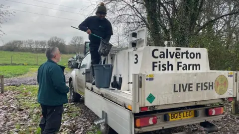 Environment Agency One man standing on the back of a van, which has a sign saying "calverton fish farm, live fish" on the back. He is standing holding a rod and net over a bucket, while another man stands on the side looking at him
