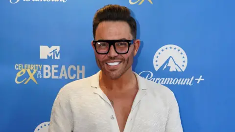 PA Media A smiling Bobby Norris looks directly at the camera, dressed in a white shirt and wearing large black-framed glasses. 