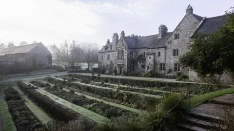 View of the front of the historic property on a misty winter's morning. The stone building has a number of small windows. A well kept rolling garden is front of the property.