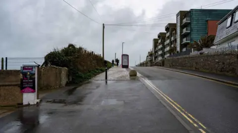 A digital screen which appears to be the size of a telephone box can be seen on the pavement at the side of the beach, with apartments seen on the other side of the road.
