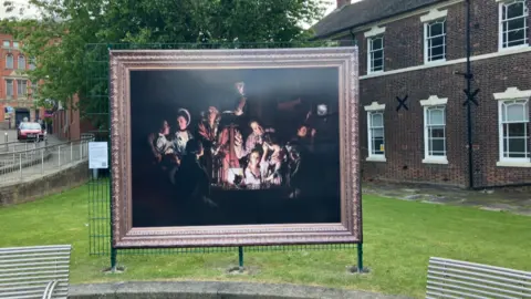 A very large reproduction of a painting, set in the grounds of a park. The painting is largely dark, and features a number of people crowded together.