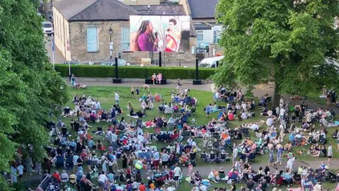 Crowd of people sat on grass looking towards a big screen, with trees on either side.