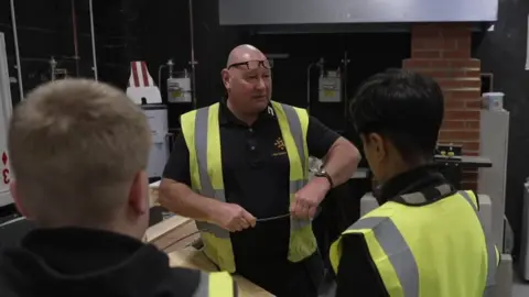 A man wearing a high visibility jacket is talking to a group of young boys, also wearing high visibility jackets. They appear to be in an industrial facility.