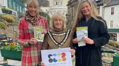 A woman wearing mayoral chains, a long beige coat and brown polo neck holds up a sign saying UK Town of Culture in front of The Old Royal Library, painted in cream with white pillars across the front. She has blonde hair. Two other women stand either side of her holding up leaflets about Malvern. Both have blonde hair, one is wearing a red and cream coat, the other a black coat.