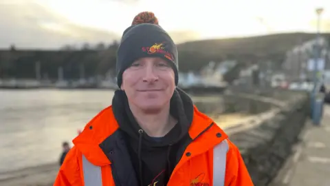 Head and upper body image of Kevin Boyle in front of the harbour at Stonehaven. He is wearing an orange hi-vis jacket over a black hoodie and a black beanie hat that says Stonehaven and has a picture of a flaming fireball 