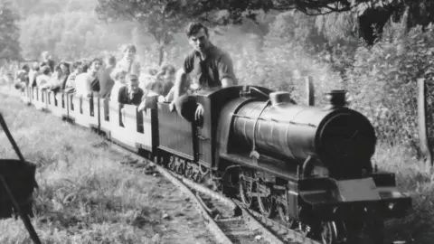 A black and white image of a miniature railway with at least seven carriages filled with parents and children and a train driver in rolled up shirt sleeves driving the miniature engine. In the background can be seen grass and trees.