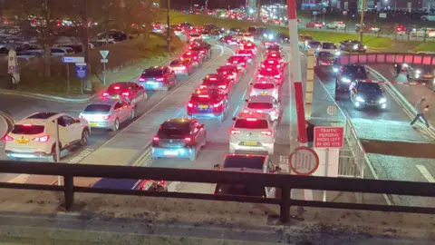 Dan Thomas Una vista desde el borde de un estacionamiento de varios pisos hacia abajo a una fila de vehículos estacionados en el tráfico, con sus luces de freno rojas iluminadas.