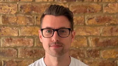 Will Lloyd-Cook smiles at the camera head on. He has short dark hair which has been pushed back. He wears a white T-shirt and black-framed glasses.