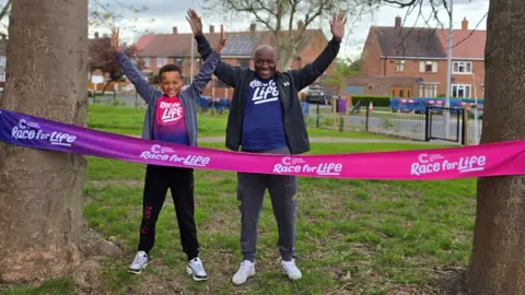 A man and a boy stand in a park with their hands in the air. They are both wearing t-shirts that say "Race for Life" on them. They are standing between two trees that have a pink banner tied across them, which also says "Race for Life" on it.