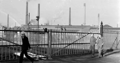 Getty Images Una foto en blanco y negro muestra a tres hombres pasando por las puertas de metal. Detrás de ellas hay varias chimeneas fumar de Corby Steelworks. 