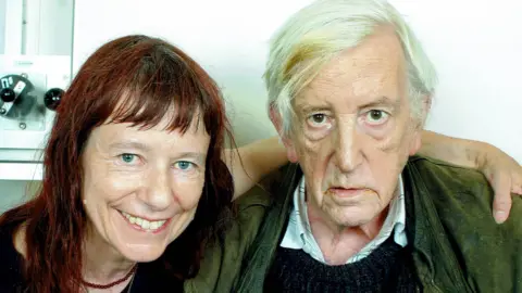 Longtime friend Rose Niland (pictured left) with long auburn hair and blue eyes with her arm around the late David Guinness who has short white hair, a long face and a dark jacket on.