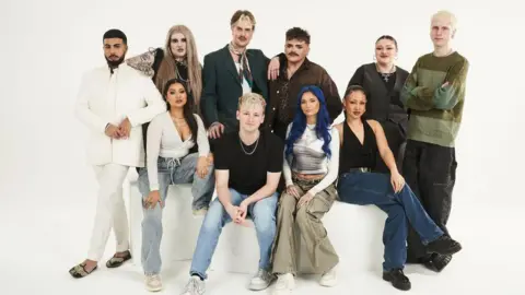 BBC/Wall To Wall/Dave King Five men and five women posting on a white bench, in a room with a white wall and a white floor.