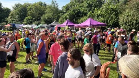 Jonathan Reed/BBC A large outdoor gathering taking place on a grassy area. In the foreground, there is a crowd of people, Many are dressed in sportswear such as running vests, shorts, and trainer. Behind the crowd, there is a row of pop-up tents in different colours.
