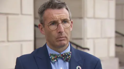 Andrew Muir standing outside Stormont. He is wearing a navy blazer, blue shirt and multi-coloured bow tie. He is wearing round glasses.
