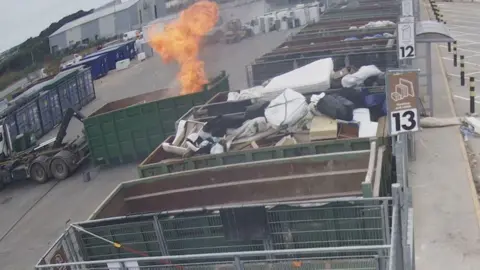 There are green skips lined up. There is a lorry reversing and pushing a green skip bin into place. There are bright orange flames coming out of the bin.