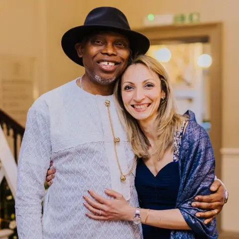 Karima Ngaage Dumbor Ngaage and his wife Karima are smiling and standing arm in arm in a hallway, in front of a staircase. Ngaage is wearing a black hat, white smock and black trousers. Karima is wearing a black evening gown and blue shawl. She is long dark blonde hair.