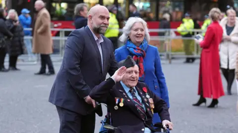 PA Media Bernard Morgan, an older man, wearing a suit and sitting in a wheelchair with a man and woman standing behind. He has numerous medals on his chest and Poppy Appeal pin badges. He is holding his right hand up in salute. The man behind him is wearing a dark-coloured suit and the woman is wearing a blue coat as well as a blue and red scarf.