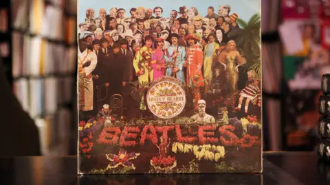 An LP cover standing on a black table against a backdrop of shelves full of vinyl albums. The cover shows the four members of The Beatles dressed in colourful military uniforms surrounded by dozens of cut-out figures of well-known people, some in colour, some black and white. The text "Sgt Pepper's Lonely Hearts Club Band" is written on a white bass drum. The word "Beatles" is spelled out in flowers.