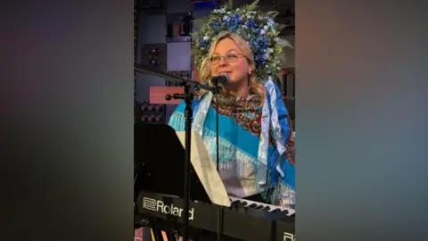 Maryna Shpylova Maryna is wearing traditional Ukrainian dress - with a blue tasselled shawl and a striking floral coronet. She is in front of a keyboard and is speaking into a mic
