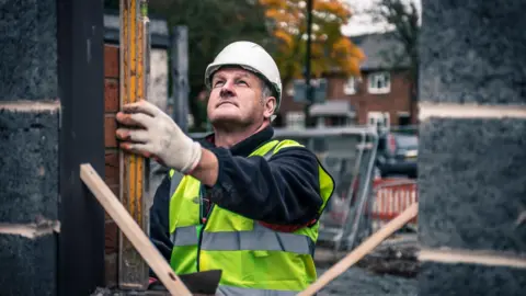 Getty Images Builder with white hat and yellow high visibility jacket, holds up a spirit level, beside brickwork