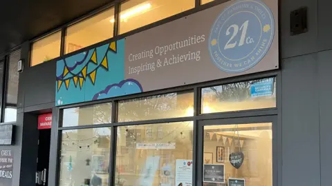 The front window of 21 Co. a cafe on Otley road in Headingley. A grey shop front with a sign above the door that reads Creating Opportunities Inspiring and Achieving. 