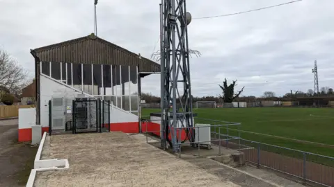 Part of the ground at Stag Meadow, with a covered terrace to the left of the picture and the grass pitch to the right, with the bottom part of a floodlight in the centre of the picture.