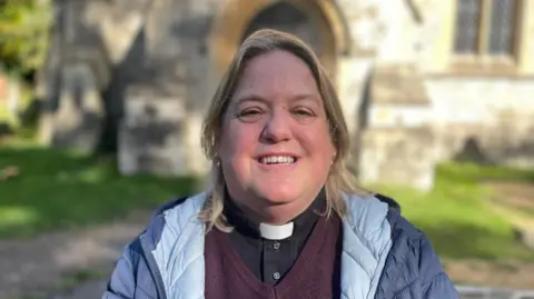 Laura Foster/BBC The Reverend Jane Yeadon stands in front of the entrance to the church on a sunny day. She has blonde hair to her shoulders and is wearing a navy blue and pale blue jacket over a purple jumper and a black shirt with a clerical collar.