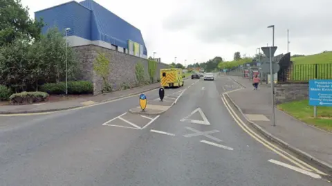 A blue building sits on top of a grey wall to the left of the picture. A yellow and red ambulance is seen heading alongside the right hand side of the building along a road. A black car is ahead of the ambulance on the road. A grey car is heading towards the ambulance on the other side of the road. 