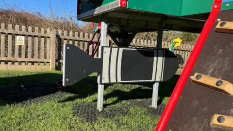 Joanna Taylor/BBC The black-and-grey shape of world war two bomb made from wood hangs below the climbing frame. Underneath is patchy grass and in the background a fence which borders the playground. 