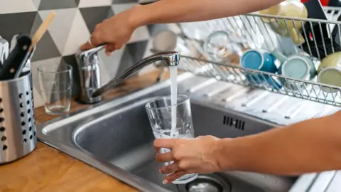 Getty Images A unidentified person filling a glass with water from a tap at a kitchen sink. Several mugs can be seen on a washing up stand in the background