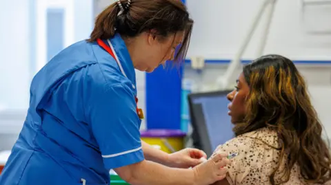A nurse in a blue uniform administering a vaccine to woman wearing a floral top in a medical setting.