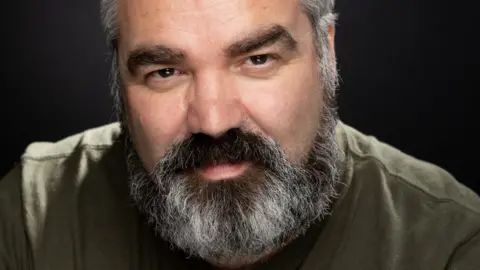 A man with a black and grey beard wearing a khaki green shirt