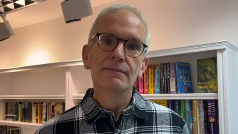 Geoffrey is looking towards the camera with a slight smile. He has black and brown round glasses, and white hair. He is wearing a blue and white checked  shirt. Behind him are shelves containing books.