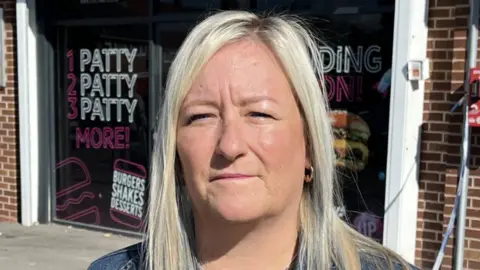 Louise is standing outside the takeaway. She has long blonde hair and a denim jacket on. The shop has black advertising signs filling the windows on either sides of the entrance. It is promoting burgers.