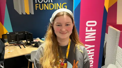 York College student Emily Goddard is looking straight into the camera. She is smiling, wearing a small, grey headscarf, a yellow lanyard and a grey T-shirt.