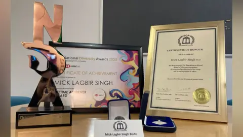 The Silver Lifetime Achiever Award is on the left of the image along with the accompanying certificate. His British Citizen Award for Volunteering and Charitable Giving is on the right, with its certificate. His table place card is in the foreground.