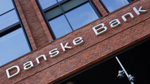 A close up of a Danske Bank sign on a red brick building. The sign is made of large white or silver letters.  The building has large windows. 