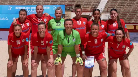 10 women stand in two rows, slightly leaning forward. The two women wearing green in the middle are flanked by 4 women wearing red kits either side. They are stood on a beach.