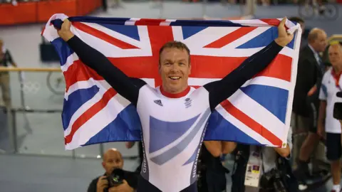 Getty Images Chris Hoy, dans sa tenue de cycliste, tient derrière lui le drapeau de l'Union après sa victoire aux Jeux olympiques de Londres 2012.