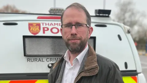 Matthew Barber is standing in a green, waxed jacket in front of a white police van.