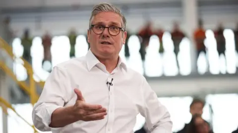 PA Media Image shows Prime Minister Sir Keir Starmer in a white shirt and glasses, speaking to workers during a visit to Glass Futures in St Helens, Merseyside, on 29 May, 2025