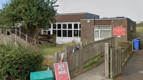 A general view of the outside of the school in Ampleforth, with views offences, trees, a pathway a blue wheelie bin, as well as the building itself with white panelled windows